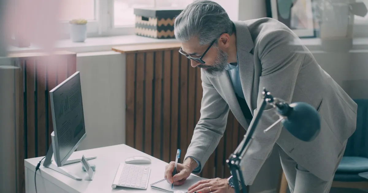 A senior compliance professional making notes at a modern office desk beside a monitor of figures