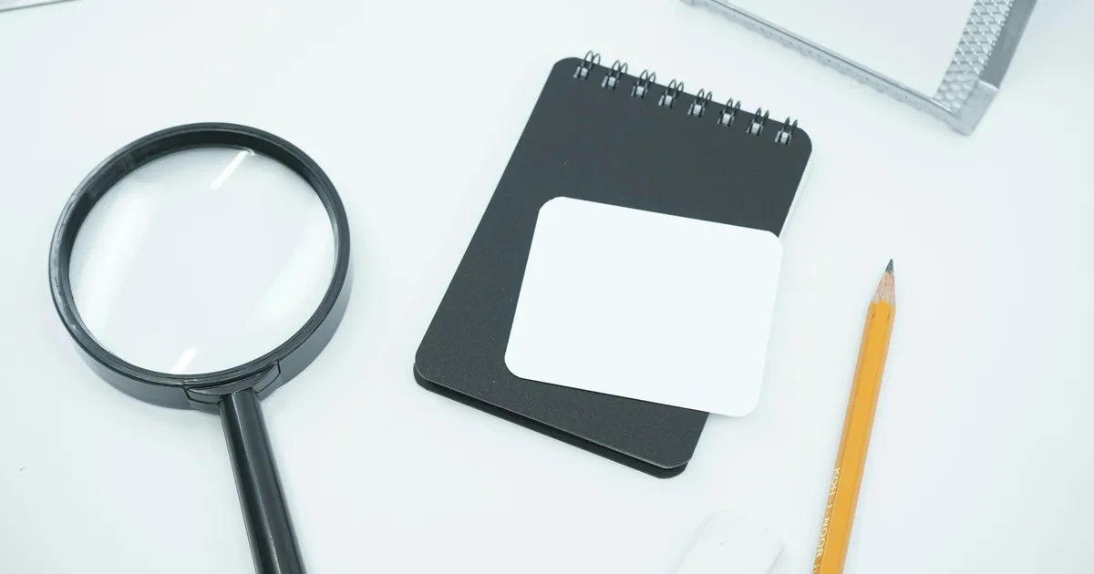 A magnifying glass, notebook and pencil on a white desk
